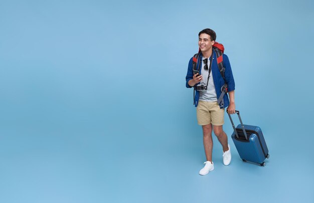 Happy young Asian tourist man holding smartphone with baggage going to travel on holidays isolated on blue copy space background