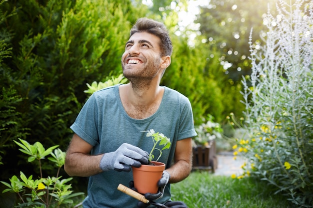 Happy young attractive bearded caucasian male gardener in blue t-shirt and gloves smiling, holding flower pot with green sprout in hands, looking aside with excited face expression