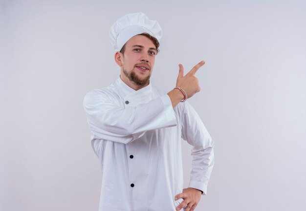 A happy young bearded chef man wearing white cooker uniform and hat pointing side while looking on a white wall