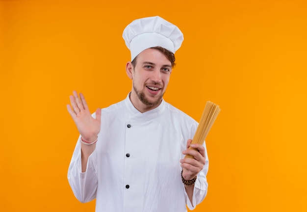 A happy young bearded chef man in white uniform holding spaghetti on an orange wall