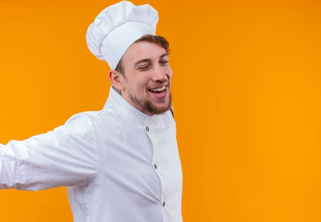 A happy young bearded chef man in white uniform opening his arms widely with close eyes on an orange wall