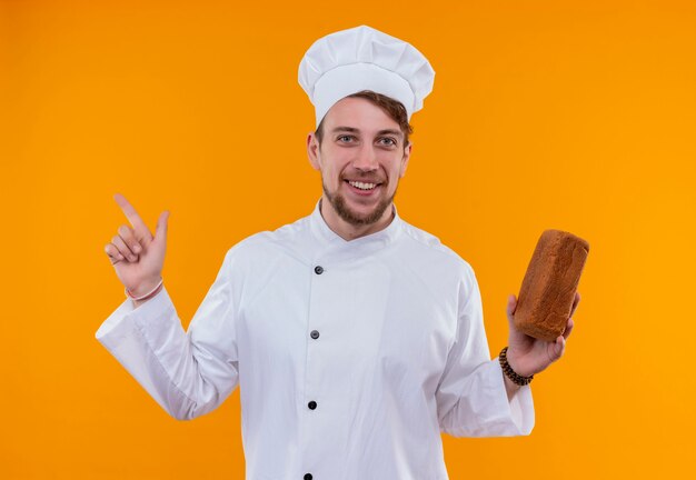 A happy young bearded chef man in white uniform pointing up with index finger while holding a loaf of bread on an orange wall