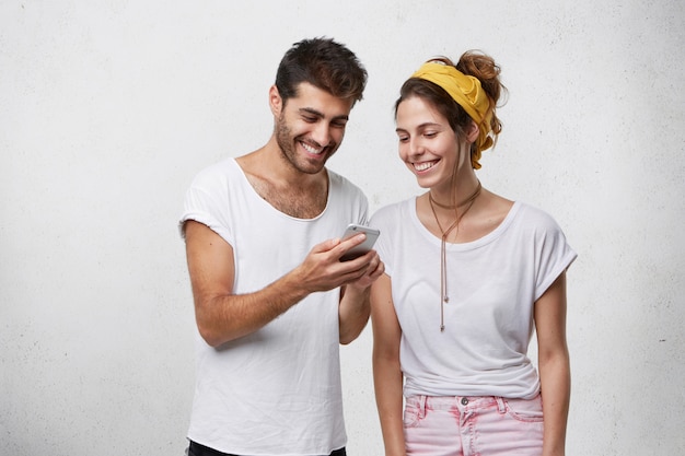 Happy young bearded hipster holding smart phone, showing his girlfriend funny pictures or videos. Relaxed carefree European couple using high-speed internet connection on electronic device together