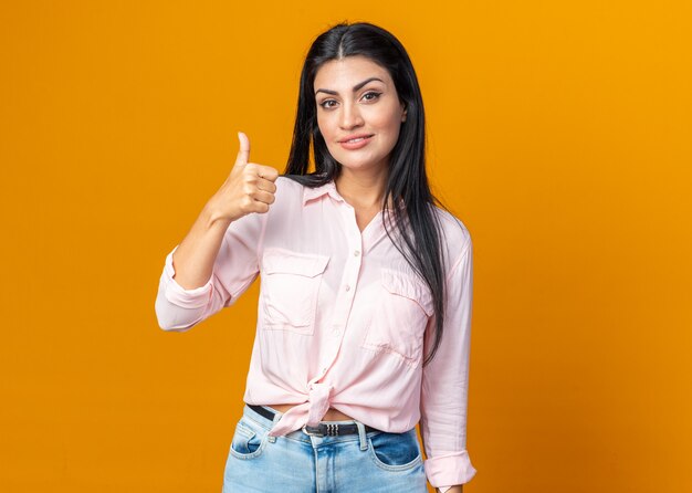 Happy young beautiful woman in casual clothes looking at front smiling confident showing thumbs up standing over orange wall