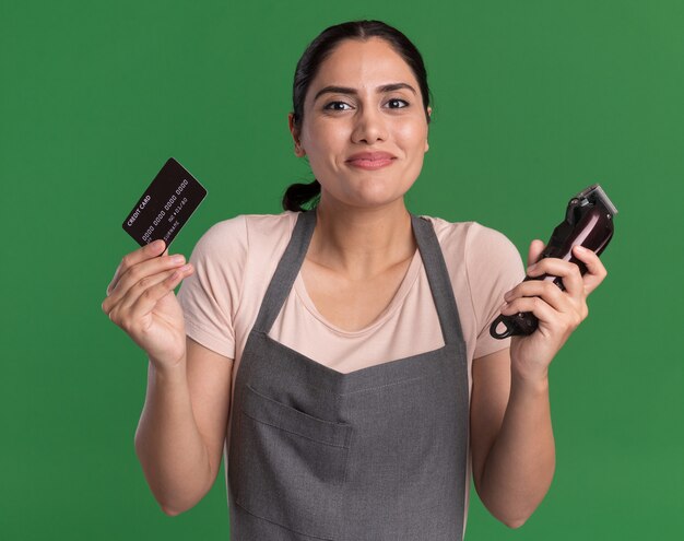 Happy young beautiful woman hairdresser in apron holding trimmer and credit card looking at front with smile on face standing over green wall