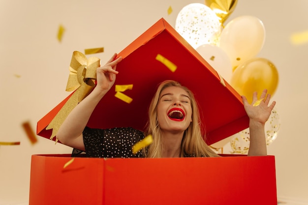 Free Photo happy young blonde caucasian girl jumps out of large gift box among confetti balloons with her eyes closed surprise concept