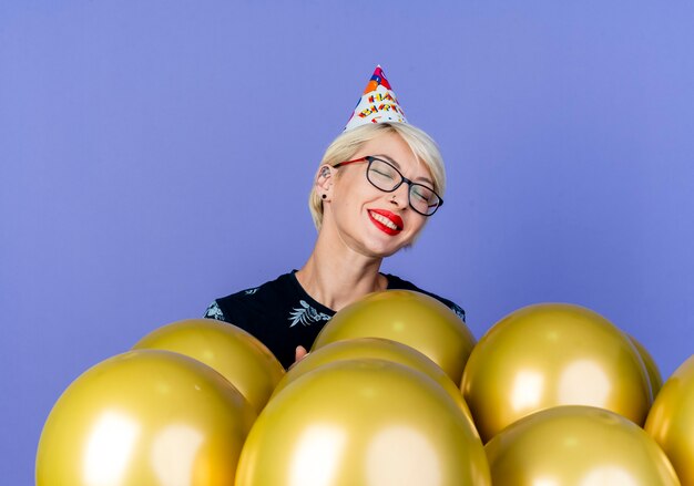 Happy young blonde party girl wearing glasses and birthday cap standing behind balloons smiling with closed eyes isolated on purple background