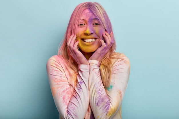 Happy young blonde woman has fun with colorful powder, keeps hands on cheeks, smiles pleasantly, expresses good emotions, entertains on Festival of Colors in spring, has painted face