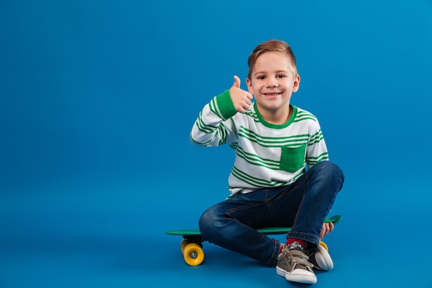 Happy young boy sitting on skateboard and showing thumb up