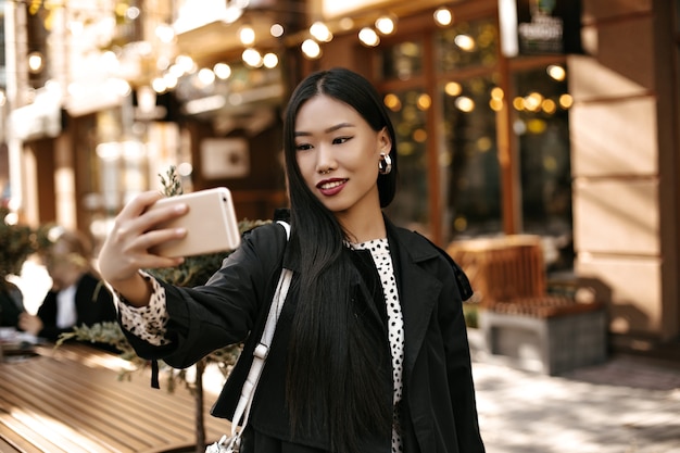 Free Photo happy young brunette lady in stylish black trench coat smiles sincerely, holds phone and takes selfie outside near street cafe