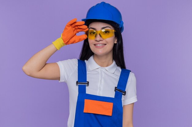 Happy young builder woman in construction uniform and safety helmet wearing rubber gloves looking at front smiling confident standing over purple wall