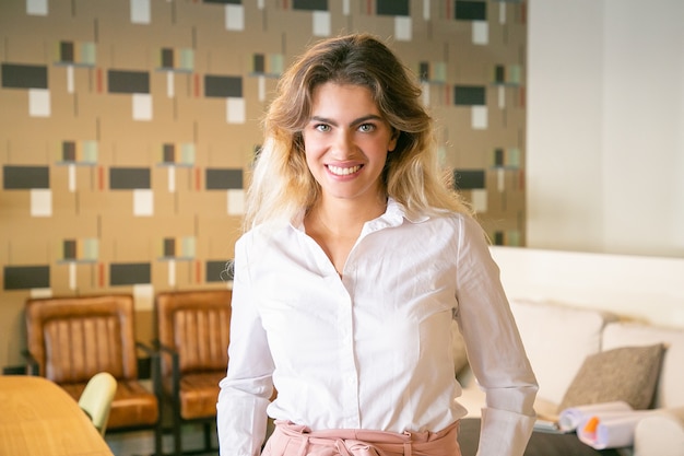 Free Photo happy young business woman standing and posing in co-working or coffee shop interior, looking at camera and smiling