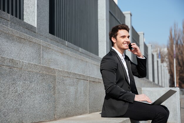 Happy young businessman sitting outdoors talking by phone.