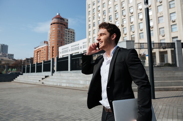 Happy young businessman walking outdoors talking by phone.