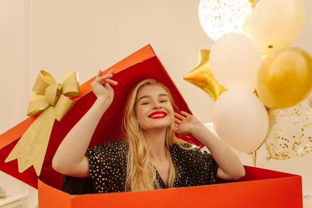 Happy young caucasian blonde woman with painted lips wears black dress while sitting in big red box People sincere emotions lifestyle concept