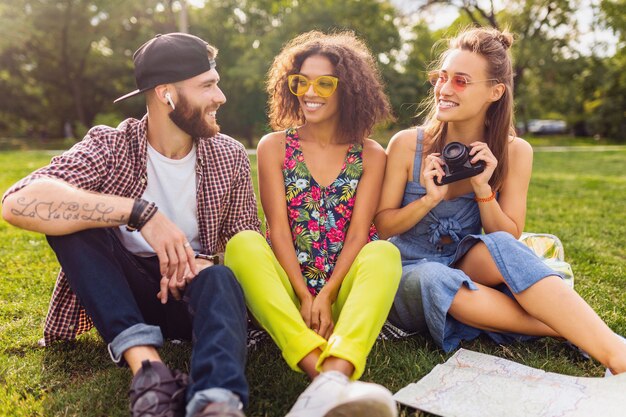 Happy young company of friends sitting park, man and women having fun together, colorful summer hipster fashion style, traveling with camera, talking, smiling