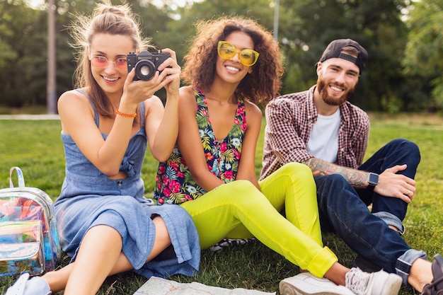 Happy young company of friends sitting park, man and women having fun together, traveling taking photo on camera, talking, smiling