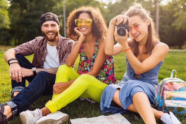 Happy young company of friends sitting park, man and women having fun together, traveling with camera, talking, smiling