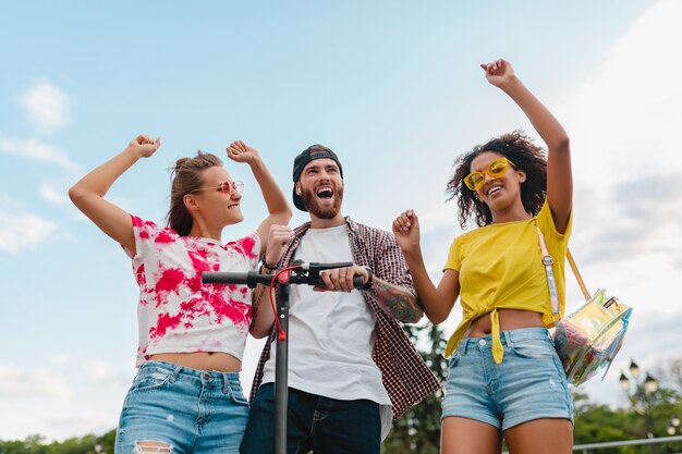 Happy young company of smiling friends dancing walking in street with electric kick scooter, man and women having fun together