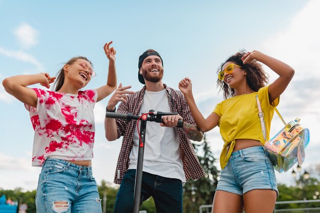 Happy young company of smiling friends dancing walking in street with electric kick scooter, man and women having fun together