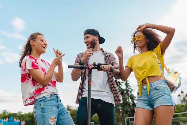 Happy young company of smiling friends dancing walking in street with electric kick scooter, man and women having fun together