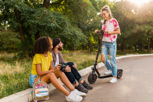 Happy young company of smiling friends sitting in park on grass with electric kick scooter, man and women having fun together