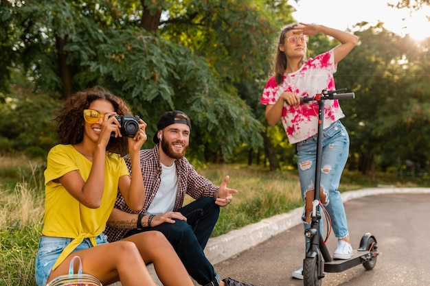 Happy young company of smiling friends sitting in park on grass with electric kick scooter, man and women having fun together