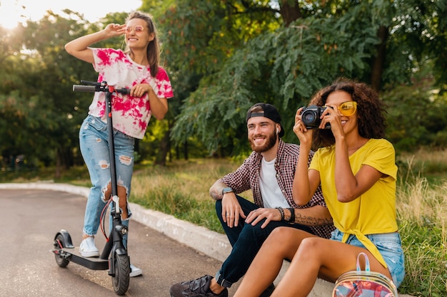 Happy young company of smiling friends sitting in park on grass with electric kick scooter, man and women having fun together