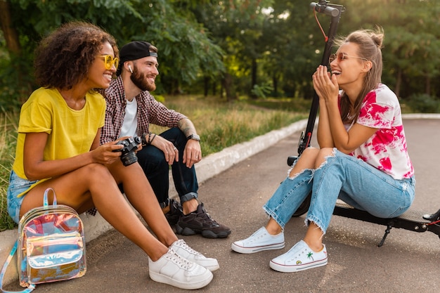 Happy young company of smiling friends sitting in park on grass with electric kick scooter, man and women having fun together