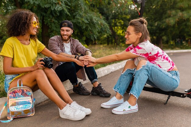 Happy young company of smiling friends sitting in park on grass with electric kick scooter, man and women having fun together