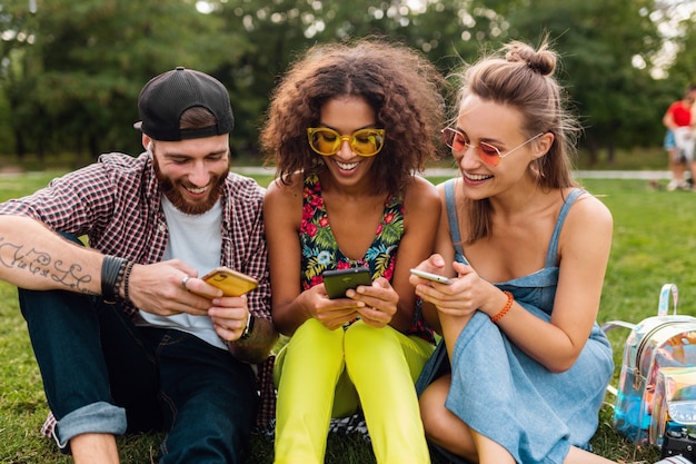 Happy young company of smiling friends sitting park using smartphones, man and women having fun together