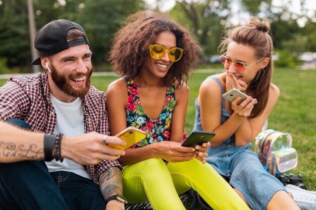 Happy young company of smiling friends sitting park using smartphones, man and women having fun together