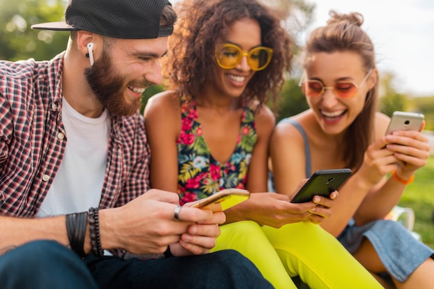 Free photo happy young company of smiling friends sitting park using smartphones, man and women having fun together