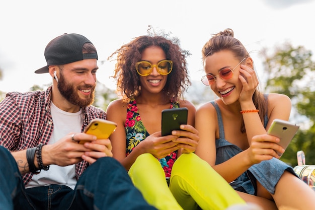 Happy young company of smiling friends sitting park using smartphones, man and women having fun together