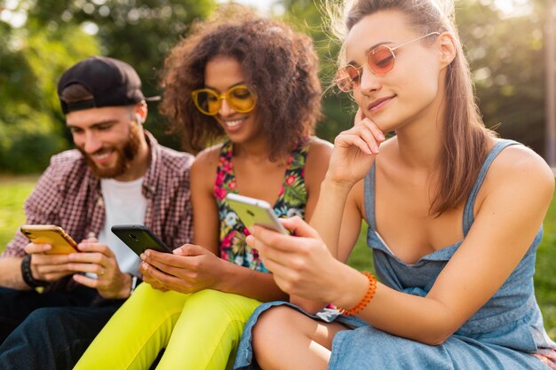 Happy young company of smiling friends sitting park using smartphones, man and women having fun together