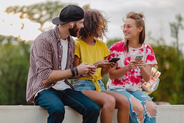 Happy young company of smiling friends sitting park using smartphones, man and women having fun