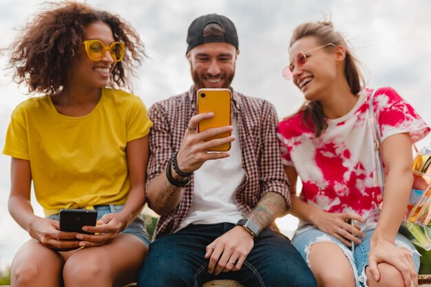 Happy young company of smiling friends sitting park using smartphones