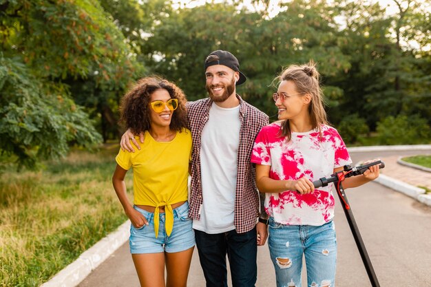 Happy young company of smiling friends walking in park with electric kick scooter, man and women having fun together