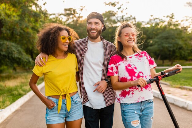 Happy young company of smiling friends walking in park with electric kick scooter, man and women having fun together