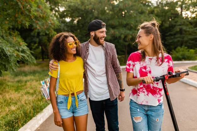 Happy young company of smiling friends walking in park with electric kick scooter, man and women having fun together