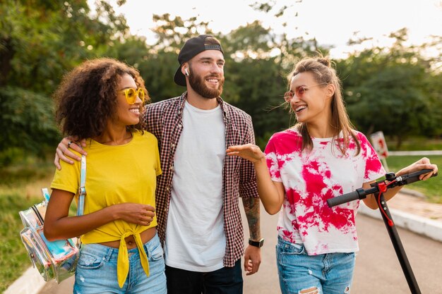 Happy young company of smiling friends walking in park with electric kick scooter, man and women having fun together