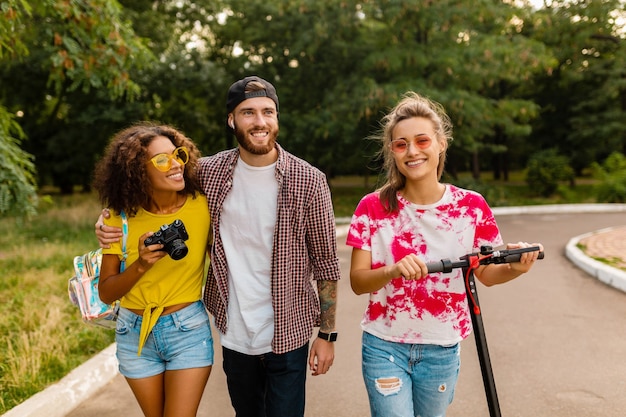 Happy young company of smiling friends walking in park with electric kick scooter, man and women having fun together