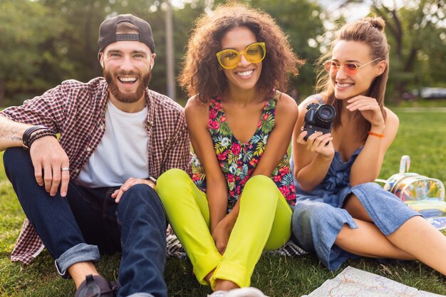 Happy young company of talking smiling friends sitting park, man and women having fun together, traveling with camera