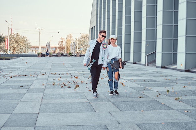 Happy young couple hold hands and walking together with skateboards on a modern street in windy weather.