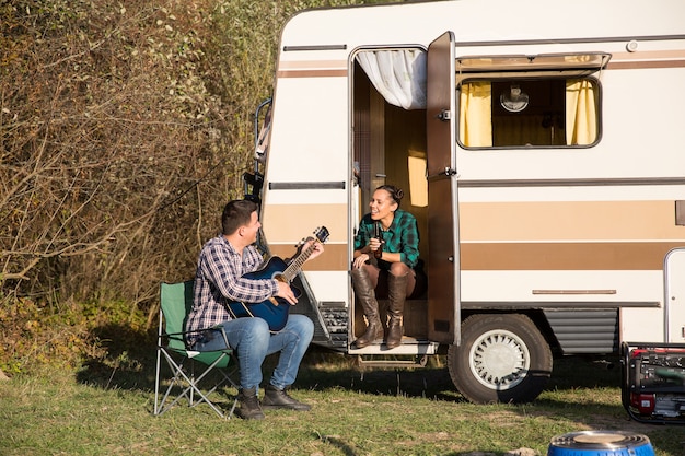 Happy young couple relaxing in the mountains with their retro camper van. Boyfriend playing on guitar for his girlfriend.