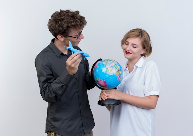 Free photo happy young couple of tourists man and woman holding globe and toy air plane standing together over white wall