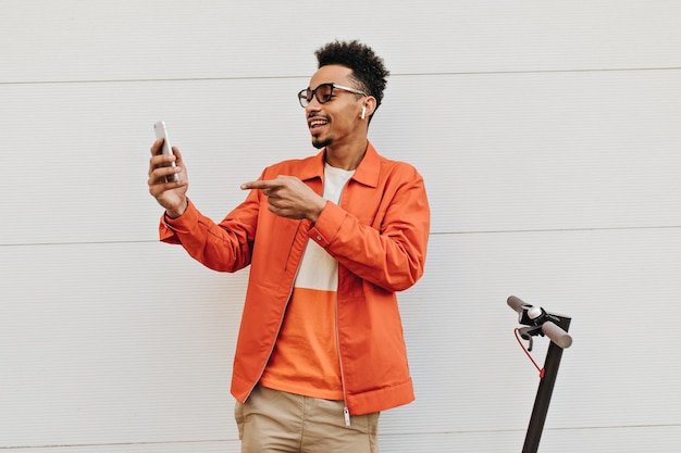 Happy young darkskinned guy in orange jacket colorful tshirt and sunglasses holds phone and talks by video outside