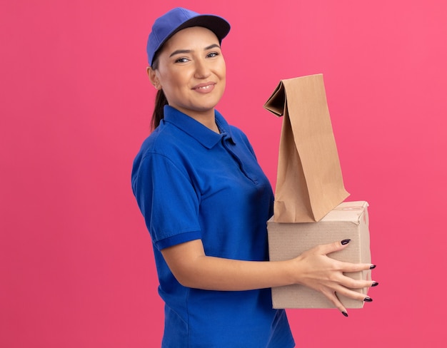 Happy young delivery woman in blue uniform and cap holding paper package and cardboard box looking at front smiling confident standing over pink wall