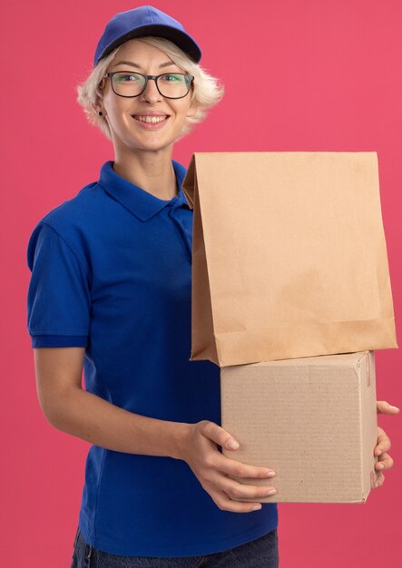 Happy young delivery woman in blue uniform and cap holding paper package and cardboard box  smiling cheerfully over pink wall