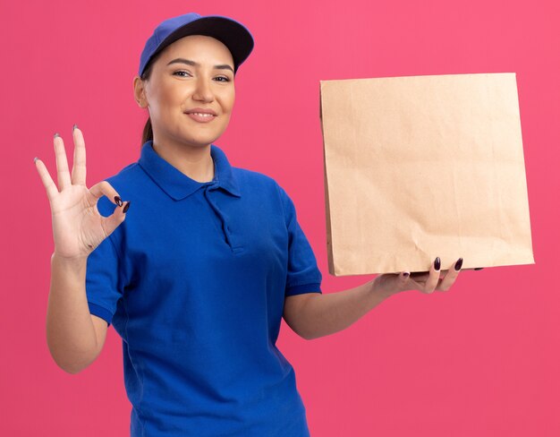 Happy young delivery woman in blue uniform and cap holding paper package looking at front smiling cheerfully showing ok sign standing over pink wall